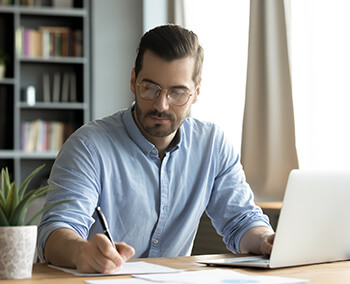 A man taking notes while checking information on his laptop